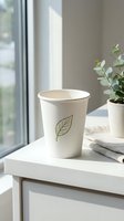Blank white disposable cup with leaf design on modern desk near window with potted plant and natural sunlight for branding and packaging mockup.