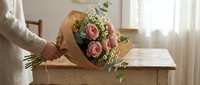 Woman holding elegant pink rose bouquet wrapped in brown paper on wooden table in bright natural light interior setting for romantic occasions.