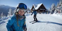 Young Caucasian girl in blue ski gear smiling on snowy mountain slope with skier and alpine lodge in background on sunny winter day.