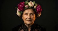 Elderly Eastern European woman wearing floral crown of pink and burgundy peonies, dark embroidered traditional costume, studio portrait on dark background.