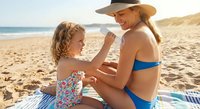 Caring mother applying sunscreen to young daughter on sunny beach during summer vacation. Family sun protection and skincare routine by ocean waves.