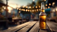 Cold beer bottle on wooden table with blurred festive outdoor party lights in background creating warm evening ambiance.