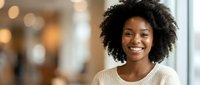Young African American woman with curly hair smiling warmly in modern office environment. Professional businesswoman portrait for corporate communications.