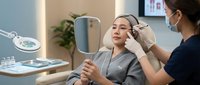 Young Caucasian woman receiving professional eyebrow treatment from Asian beautician in modern cosmetic salon with magnifying lamp and mirror.