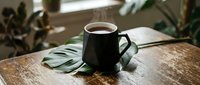 Dark ceramic coffee mug with hot beverage on rustic wooden table surrounded by green tropical leaves in bright natural window light for cozy morning routine.