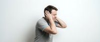 Young Caucasian man in gray t-shirt covering ears with hands, expressing stress or avoiding noise against white background for mental health concepts.