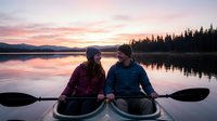 Couple in kayak on calm lake at sunset with forested mountains and pink sky reflected in water, outdoor recreation and adventure activity.