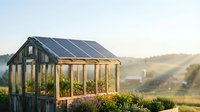 A wooden greenhouse with solar panels on the roof sits in a rural landscape during golden hour, surrounded by flowering plants and countryside views, suitable for sustainable agriculture and eco-friendly living concepts.