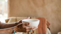 Mature man with brown skin holding empty white cup with both hands, wearing casual clothing and turquoise bracelet, indoor setting with warm neutral background for charity, donation, or help concept.
