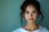 Young Asian woman with natural makeup and messy bun hairstyle wearing white t-shirt against turquoise background, soft lighting creates gentle mood and highlights facial features.