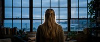 Young woman with long blonde hair sitting at desk looking through large industrial windows at city skyline during evening hours in modern office workspace.