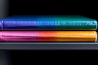 Colorful stack of hardcover notebooks with rainbow gradient spines against dark background, showing vibrant transition from blue to turquoise and pink to orange.