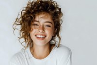 Happy young woman with curly brown hair smiling broadly in white t-shirt against light background. Natural portrait for lifestyle and wellness content.