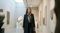 Woman with shoulder-length brown hair, Caucasian ethnicity, wearing dark coat and patterned scarf, walking through contemporary art gallery with white walls, sculptures and framed artwork displayed. Suitable for art, culture, museum, and lifestyle concepts.