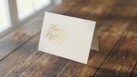Elegant cream greeting card with gold foil Happy Birthday text on rustic wooden table surface for celebration and party invitations.