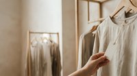 Caucasian female hand touching linen garment on wooden hanger in minimalist closet with neutral tones, suitable for fashion, lifestyle, and home organization content.