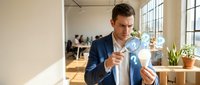 Caucasian businessman in blue suit examining documents with magnifying glass in modern office workspace for business analysis and investigation.