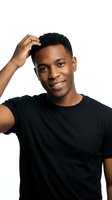 Young African American male with short textured hair, wearing black casual t-shirt, touching head with hand while smiling at camera against white background for portrait and lifestyle use.