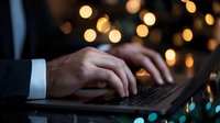 Male hands typing on laptop keyboard in dark room with blurred bokeh lights background, creating atmospheric evening work environment.