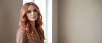 Young redhead woman with wavy hair in beige blazer looking thoughtfully away in modern office interior with natural window light for business portraits.