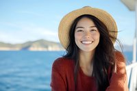 Young Asian woman wearing straw hat and red sweater smiling brightly while enjoying boat trip on blue sea, wind blowing through long dark hair, mountains in background.