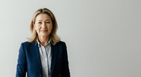 Middle-aged woman with shoulder-length blonde hair, Caucasian ethnicity, wearing dark navy blazer over light blue shirt, professional business portrait against neutral gray background.