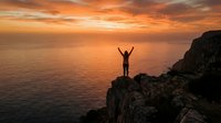 Person standing on rocky cliff with arms raised at sunset over ocean, celebrating achievement and freedom during golden hour.