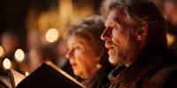 Senior Caucasian man with gray beard singing Christmas carols by candlelight with choir group during evening holiday celebration in warm atmospheric setting.