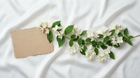 Blank kraft paper card with white jasmine flowers and green leaves on white fabric background, suitable for wedding invitations, greeting cards, and stationery design.