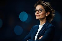 Confident middle-aged businesswoman wearing glasses and navy blazer with arms crossed, smiling against dark background with blue bokeh lights. Professional portrait.
