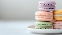 Colorful French macarons in pastel purple, peach, and mint green shades stacked on white plate against light background, showcasing delicate meringue texture and filling.