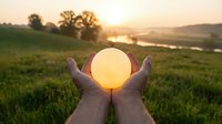 Hands holding glowing orb at sunset over green field and river landscape, symbolizing hope, energy, and nature connection for wellness and environmental concepts.