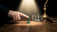 Businessman in dark suit moving green chess piece on wooden board during strategic game with brass lamp and books in background.