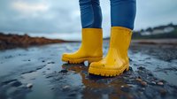 Person wearing bright yellow rubber boots standing in shallow water on wet beach sand with cloudy sky background for outdoor adventure and rainy weather concepts.