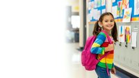 Happy Hispanic elementary school girl with pink backpack standing in bright classroom hallway ready for learning adventure.