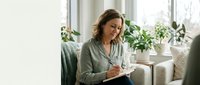 Middle-aged Caucasian woman writing in notebook while sitting on couch at home with plants by window. Comfortable indoor lifestyle and productivity concept.