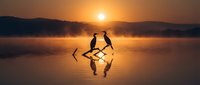 Silhouetted figures stand on shallow water at sunrise, holding oars, with golden light reflecting on the surface and misty mountains in the background. Perfect for travel and adventure themes.