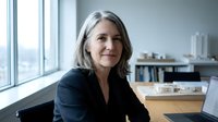 Mature woman with gray hair wearing dark blazer sitting at desk in modern office with city view and bookshelves, professional portrait for business and corporate use.