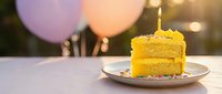 Yellow birthday cake slice with lit candle on plate, colorful balloons in background creating festive celebration atmosphere for party events.