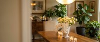 Cozy dining room interior with wooden table, fresh flowers in vase, green plants and warm ambient lighting creating inviting home atmosphere.
