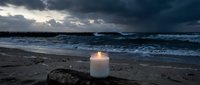 Glowing white candle on sandy beach at twilight with dramatic stormy clouds and ocean waves creating peaceful meditation atmosphere for relaxation concepts.