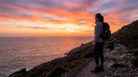 Young woman with dark hair, wearing athletic backpack and dark clothing, standing on rocky coastal cliff overlooking ocean during golden sunset. Adventure travel and hiking concept.