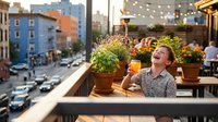 Young woman enjoying morning coffee on sunny urban balcony with colorful flowers and string lights overlooking busy city street below.