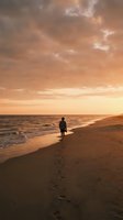Silhouetted male surfer holding board walking along sandy beach at golden hour sunset with footprints trailing behind, ocean waves and dramatic cloudy sky for travel and lifestyle content.