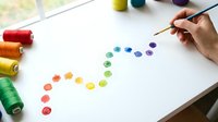 Hand painting colorful watercolor dots in rainbow spectrum on white paper with brush and thread spools nearby for creative art projects and design work.