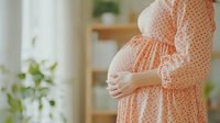 Pregnant woman in peach polka dot dress tenderly holding baby bump, standing by window with soft natural light and green plants, creating peaceful maternal moment.