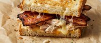 Grilled sandwich with crispy bacon strips and melted cheese on toasted artisan bread, served on parchment paper for comfort food and cafe menu concepts.