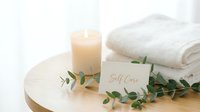 Soft spa setting with glowing candle, folded white towels, eucalyptus branch, and self care card on wooden table for wellness concept.
