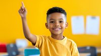 Young African American boy in yellow t-shirt raising hand enthusiastically against vibrant yellow wall, expressing excitement and engagement in classroom setting.