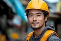 Young Asian male construction worker wearing yellow hard hat and safety vest smiles confidently at construction site, displaying professional expertise and workplace safety.